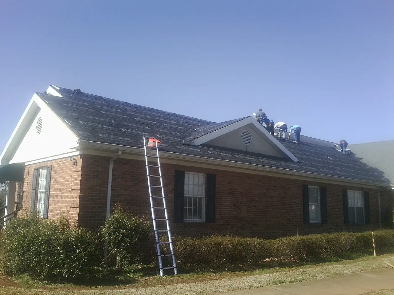 Crew tearing shingles off a brick ranch roof with a ladder set — Emergency Roof Repair work by our crew in Pfeifer