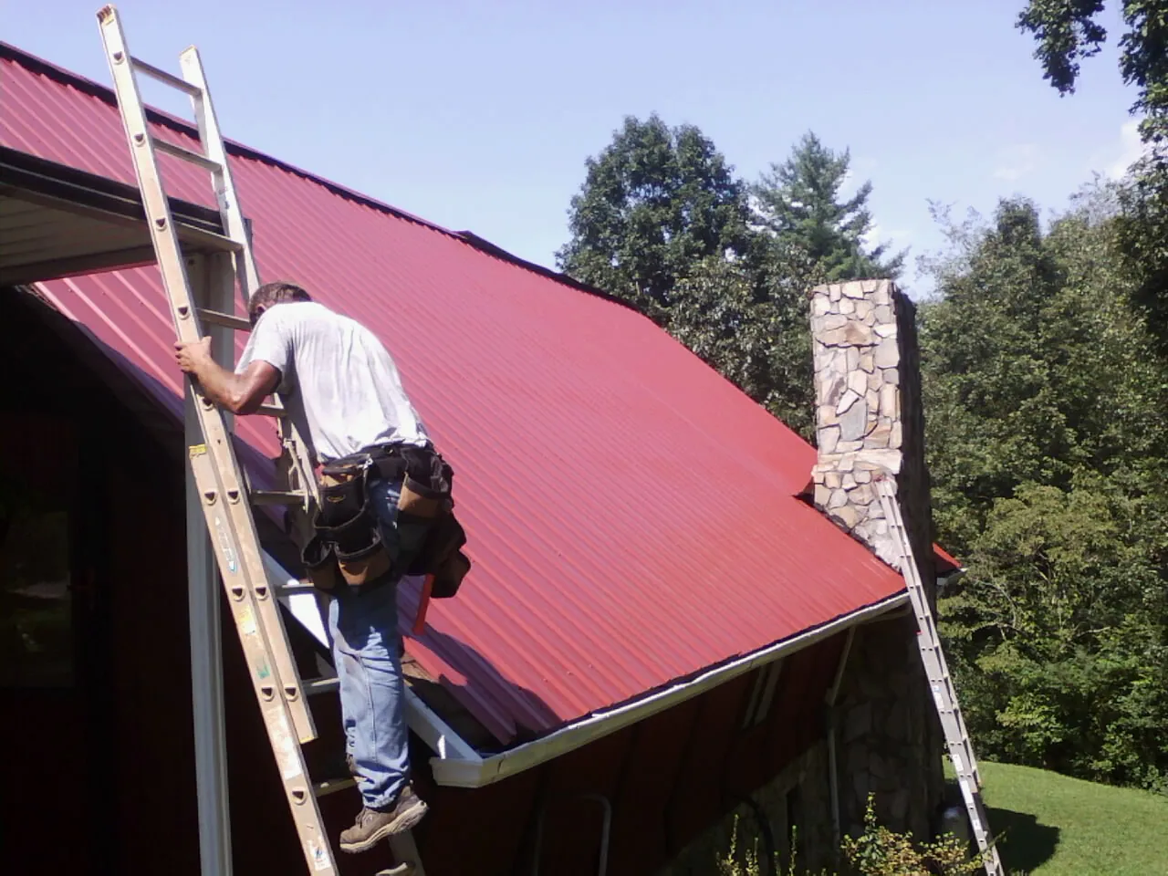 Roofer on a ladder beside a red metal roof and stone chimney — Soffit & Fascia Repair by Roofing Brothers in Pfeifer